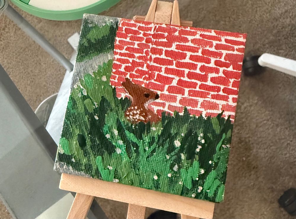 A tiny canvas with an acrylic painting of the mentioned photo; it shows a fawn sitting beanlike in front of a brick wall in the tall grass with some gravel roads to the side.