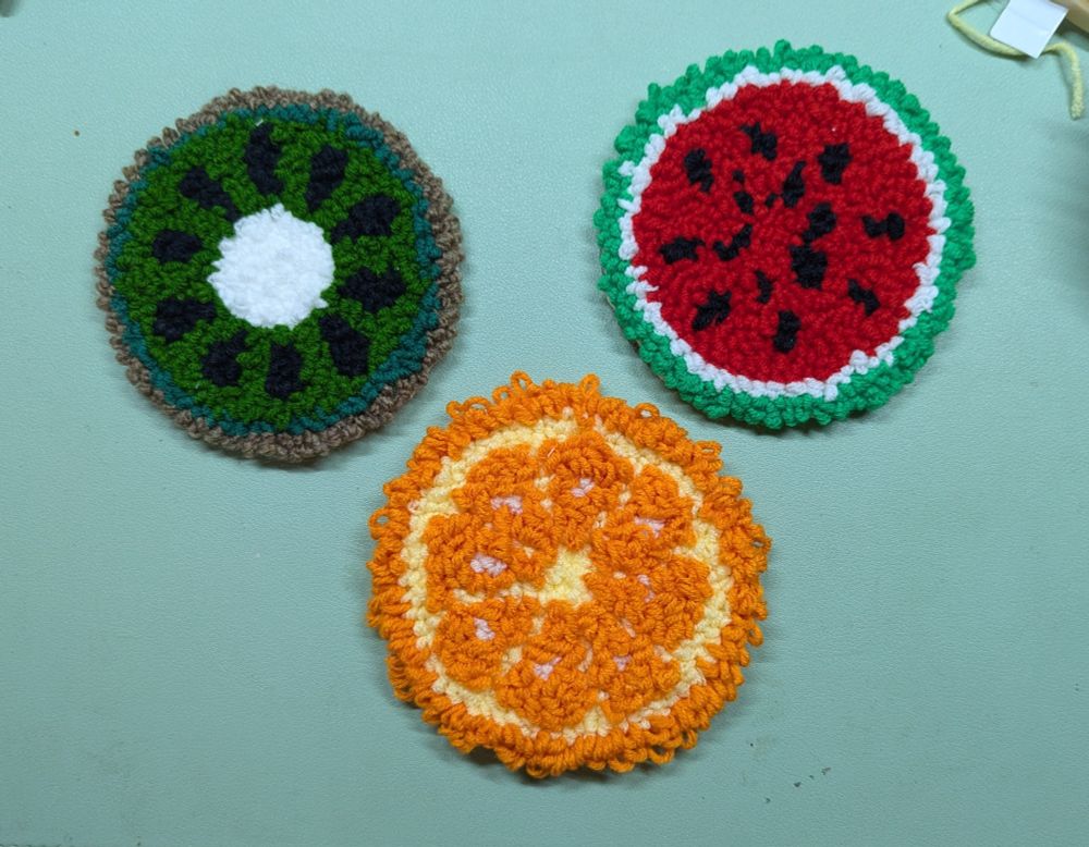 Draufsicht Foto von drei Untersetzern mit Frucht Design. Sie sind noppig wie ein Teppich

Eine Kiwi Scheibe
eine Wassermelonenscheibe
eine Orangenscheibe mit sichtbar unterschiedlicher Noppigkeit. Teile der Frucht sind höher bzw niedriger als der Rest

Alle drei sind mit einer Punch Needle "gestickt". Die Untersetzer haben eine Filzrückseite, die jedoch im Foto nicht zu sehen ist