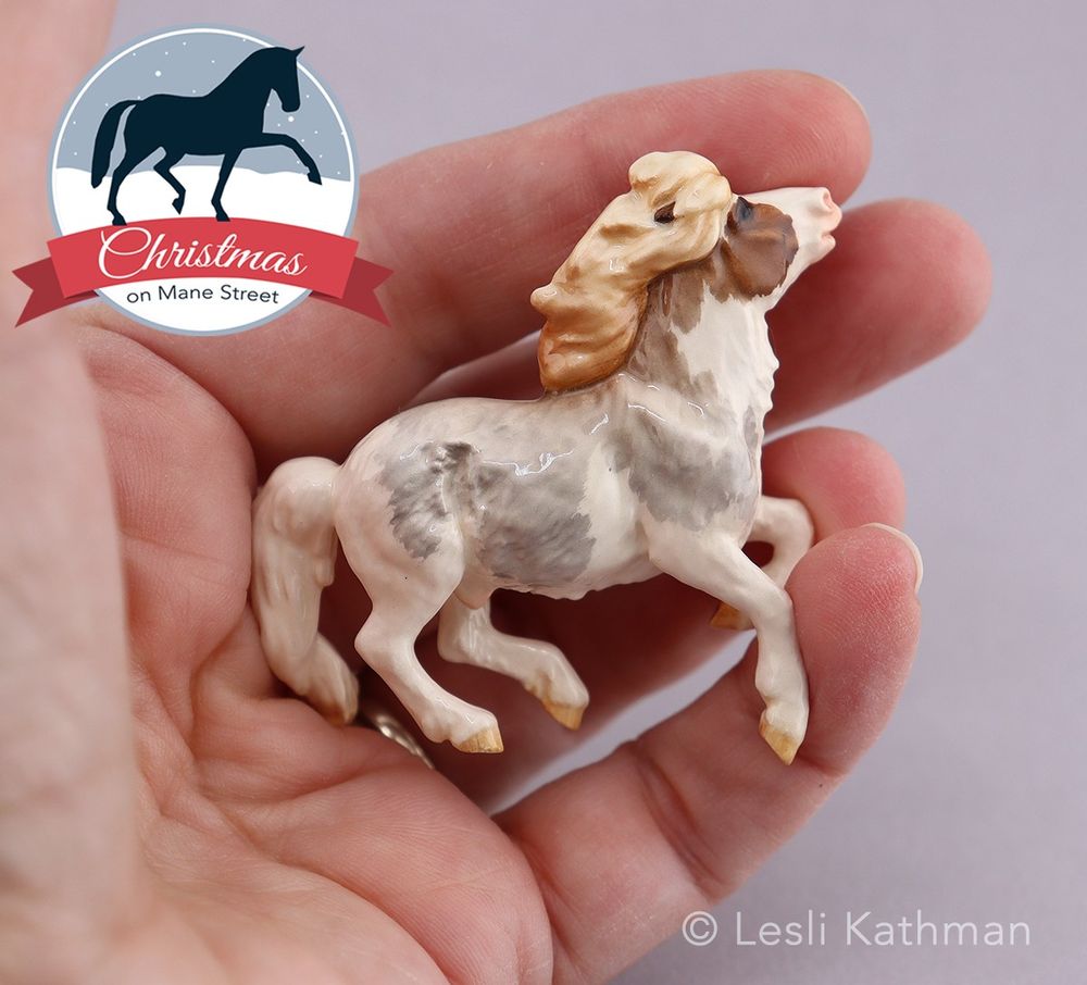 A ceramic Shetland Pony in liver roan splashed white is pictured in the artist's hand, showing the scale (approximately 2" tall) 
