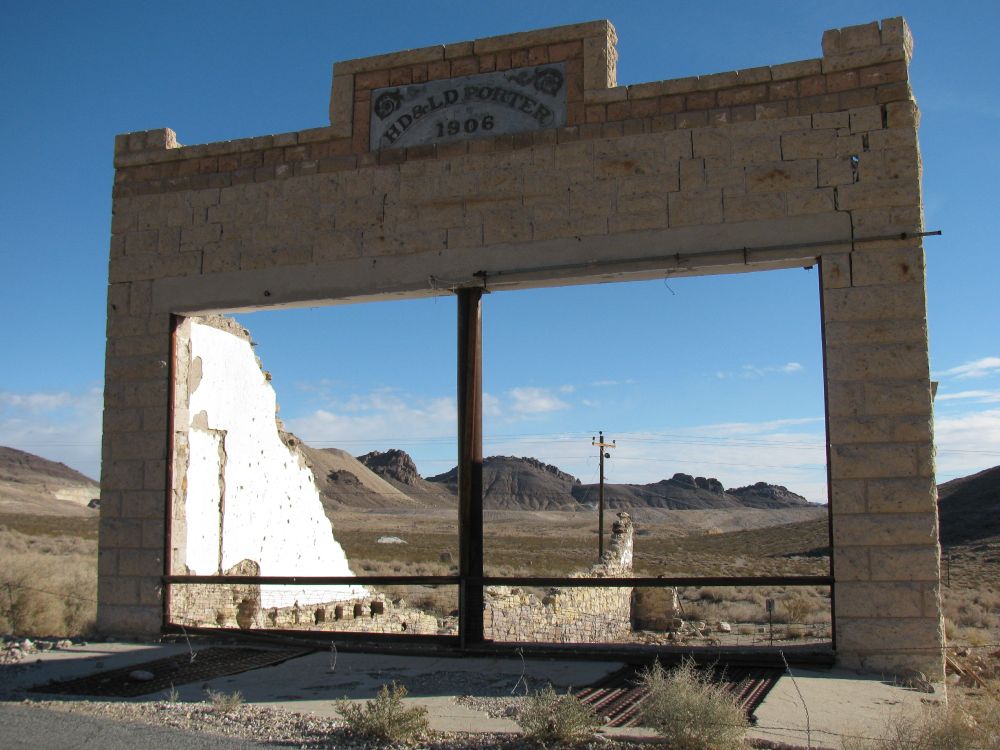 Ruine d'un ancien magasin général. On peut voir le paysage désertique  à travers l'ouverture des anciennes vitrines. Un poteau supportant des fils électriques est visible. Le ciel est bleu. Rhyolite, ville fantôme au Nevada. (2009).