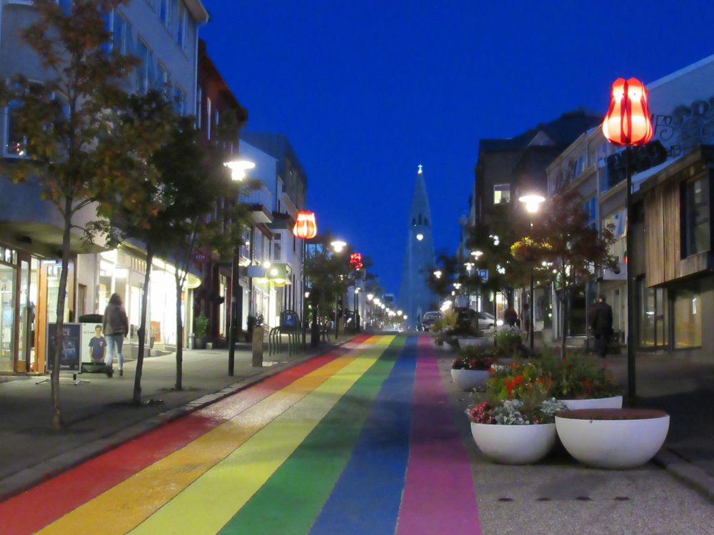 Une rue, sur laquelle est peint un arc-en-ciel, mène à l'église Hallgrímskirkja. De chaque côté de la rue, on retrouve des commerces. Le ciel est d'un bleu vibrant. Reykjavik, Islande (septembre 2019)