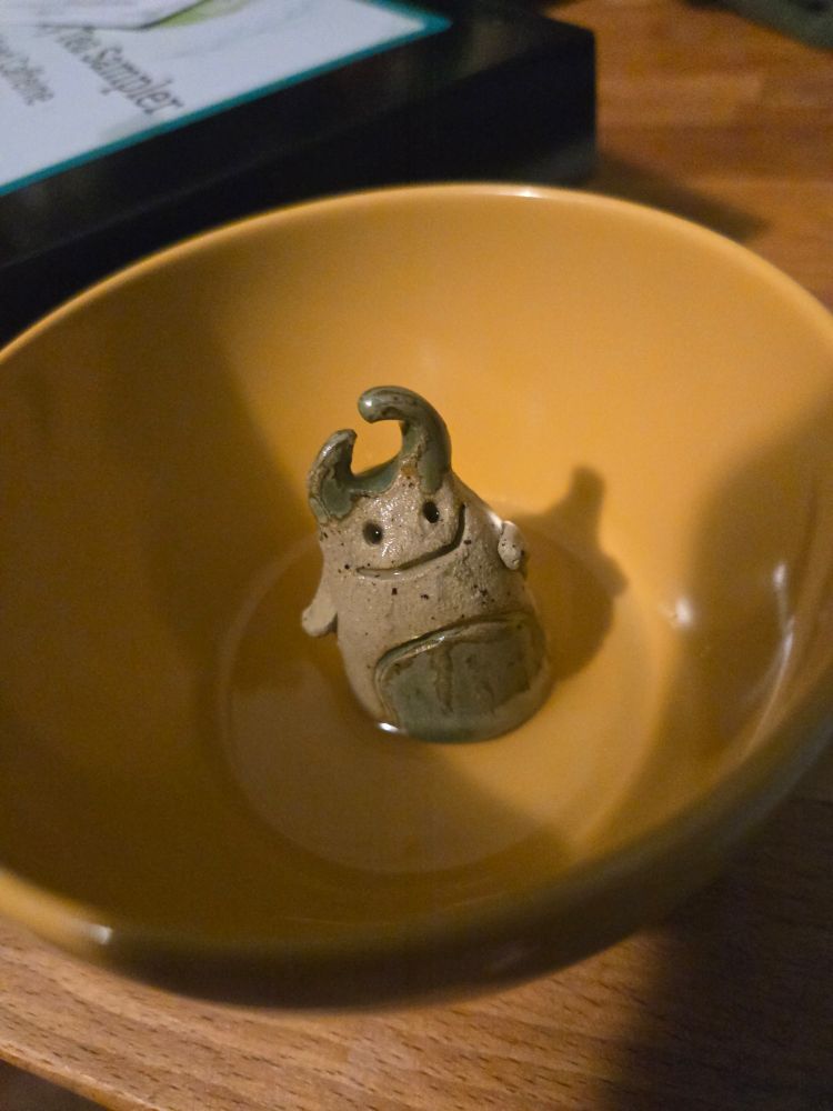 A photo showing my new ceramic "tea pet" after his first tea time. A small figure with a wide smile and two crooked horns sits in a yellow tea cup, with a shallow pour of tea surrounding him.