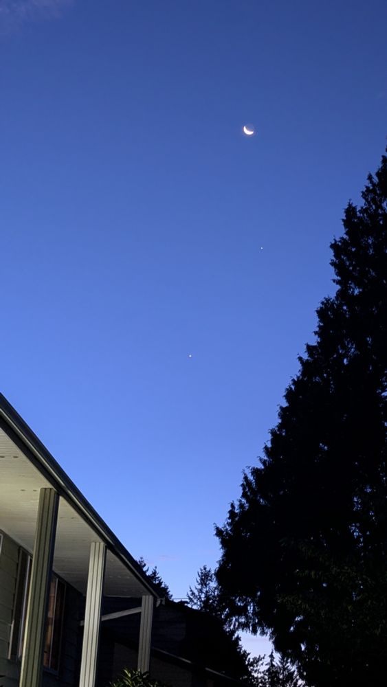 Wide view of sky, blue just before sunrise. A waning crescent moon overhead, then two smaller bright dots of light beneath them. 