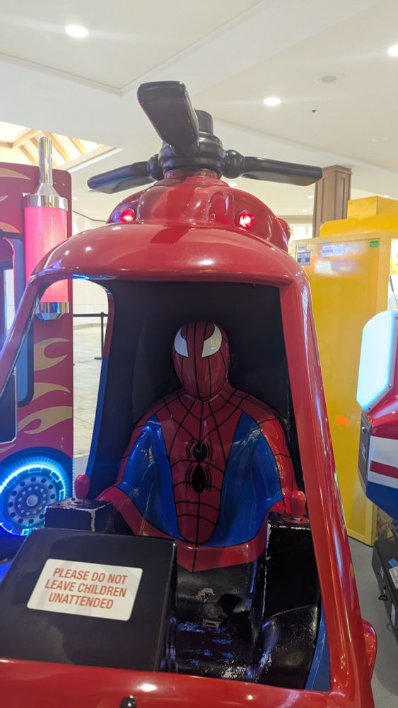 A photo of a shopping centre ride shaped like a helicopter with a very off-model Spider-Man forming the pilot seat. 