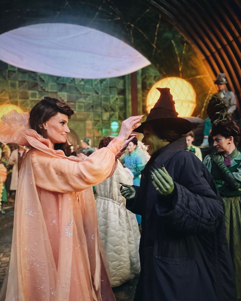 Idina Menzel dressed in a peach-colored gown adjusting the witch hat of a green-skin-painted Cynthia Erivo on the set of the Wicked film.
