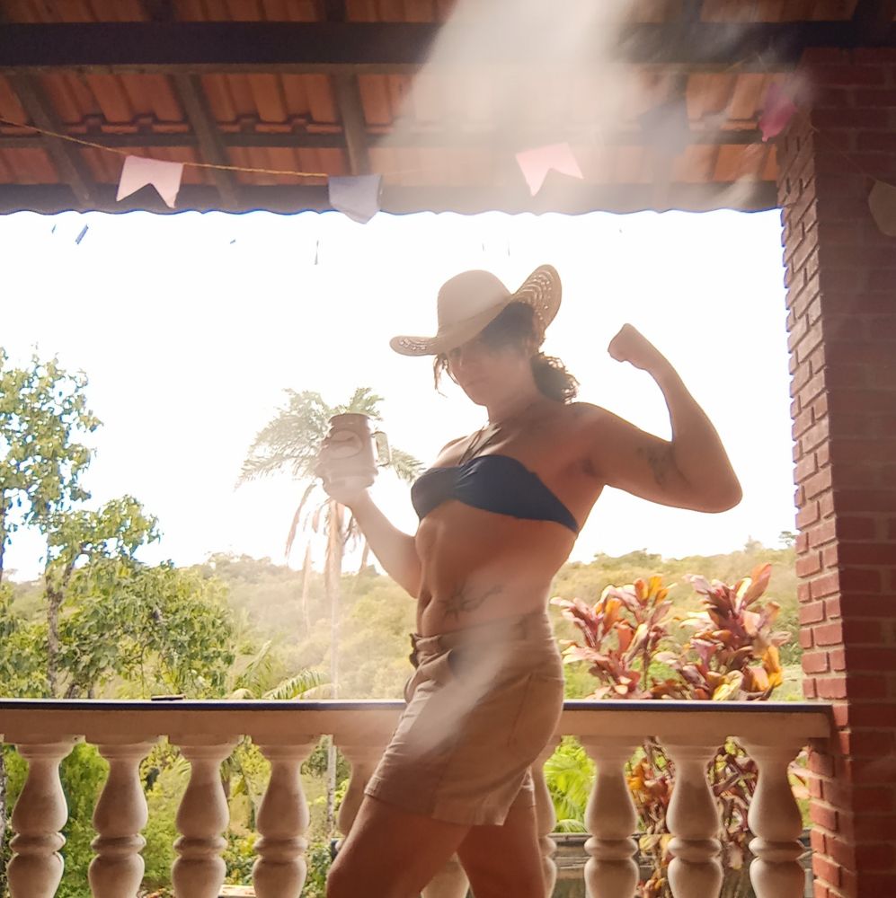 Foto minha com biquíni azul, shorts, chapéu de cowboy e uma caneca na mão 