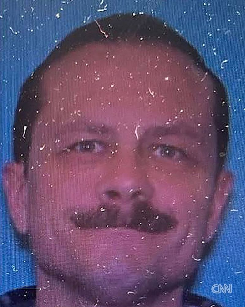 A close-up photograph of a man with short, dark hair and a prominent mustache is displayed. He is smiling slightly, positioned against a blue background. His complexion is fair, and he has a subtle shadow on his jawline. There is some visual noise and graininess in the image, obscuring parts of the man's features. His eyes are directed towards the camera, giving a direct and relaxed expression. The image signifies a police identification or mugshot context.