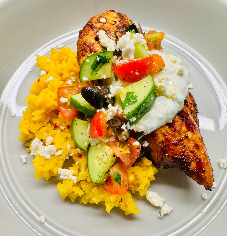 Grilled lemon harissa chicken over saffron rice and topped with tzatziki, tomato and cucumber salad and feta