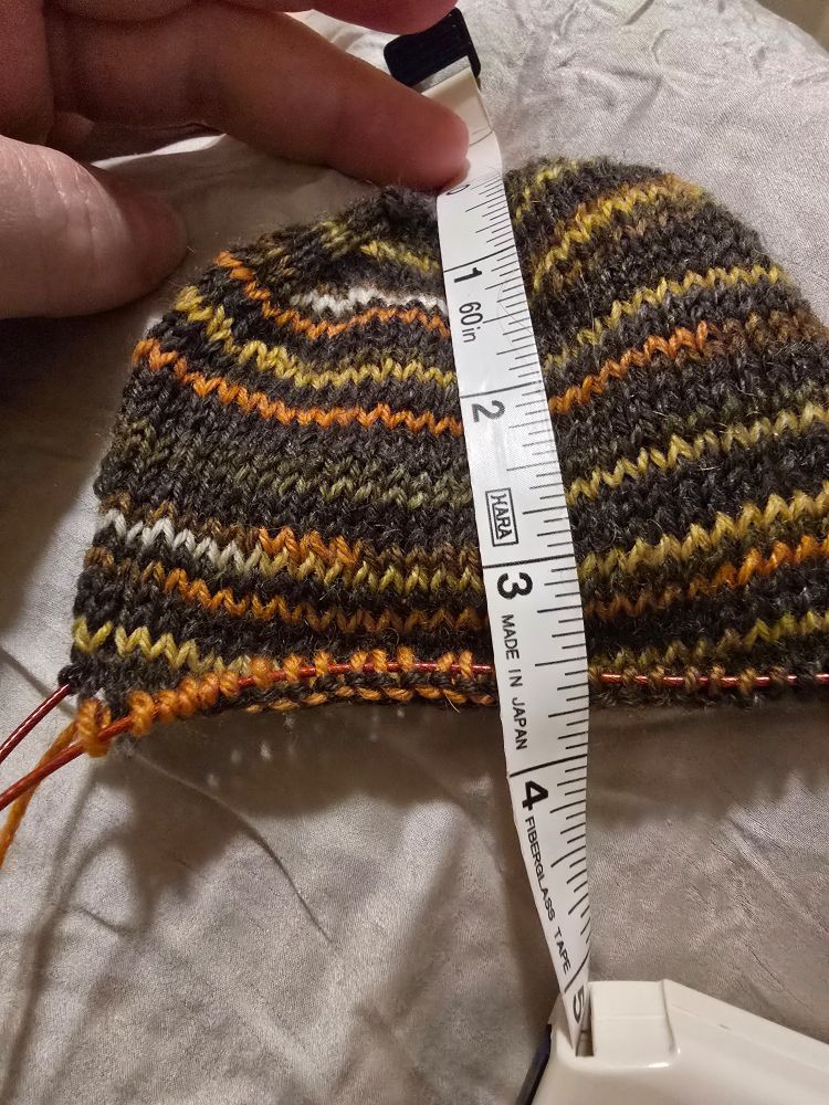 A tape measure atop 3.5 inches of toe-up handknit sock in self striping yarn. Main color is a charcoal gray, stripes are orange, yellow, and white. 