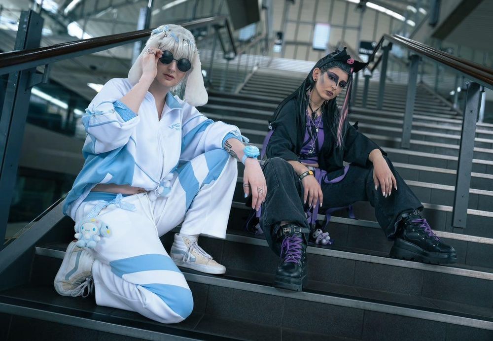 Two people sit on the stairs, on in a blue and white tracksuit, white wig and ears. The other person is in black and purple streetware, Gothic makeup and black wig