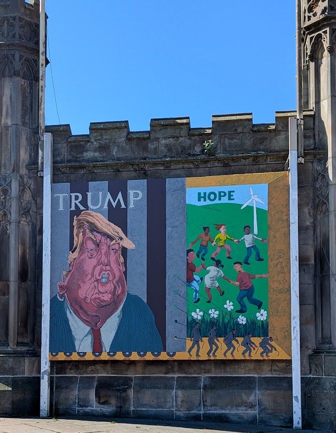 street art on the side of an Edinburgh church.  on one side "Trump", with his ugly face against a grey and black striped background, and beside it "Hope" showing children playing on a green hillside with flowers and a wind turbine. 