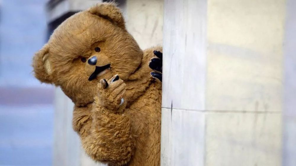 Photograph of Bungle, the bear from the kids’ TV programme Rainbow, peering out from behind a pillar and pointing. 