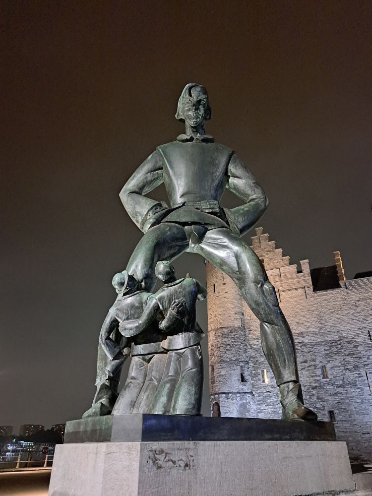 Statue of giant with legs apart and two smaller figures looking up at him. 