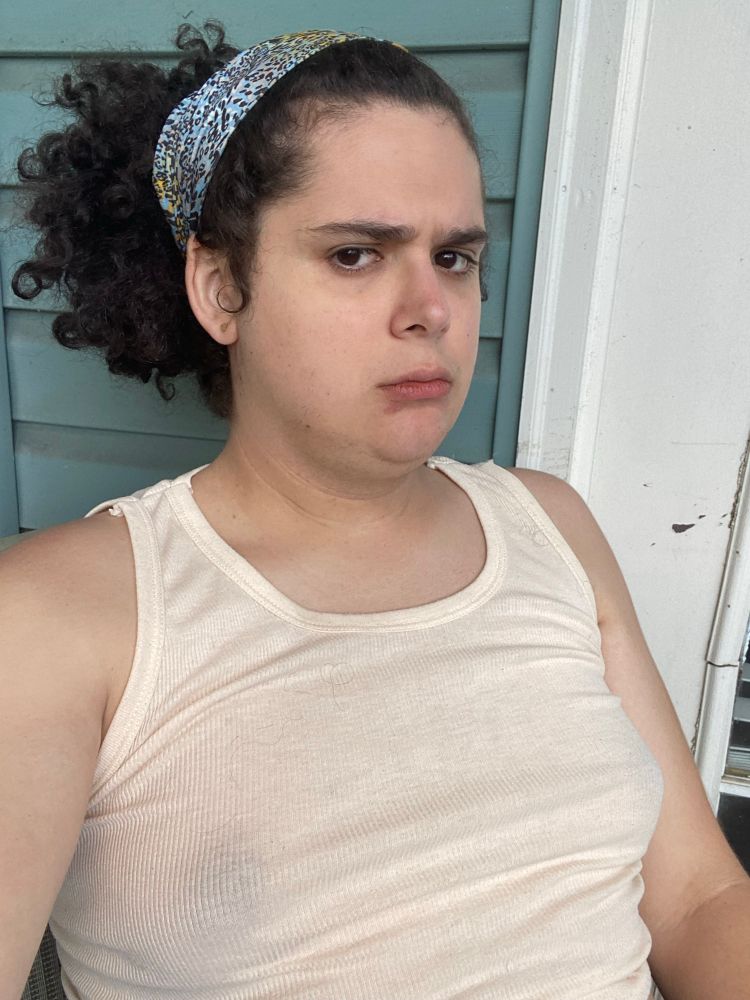 Kara is sitting on her porch with her hair in a bandana, making one of those attractive pouty faces that are all the rage these days. She is wearing a cream colored tank top with her dark areolae clearly visible on top of boobs that she would personally consider “bodacious”.
