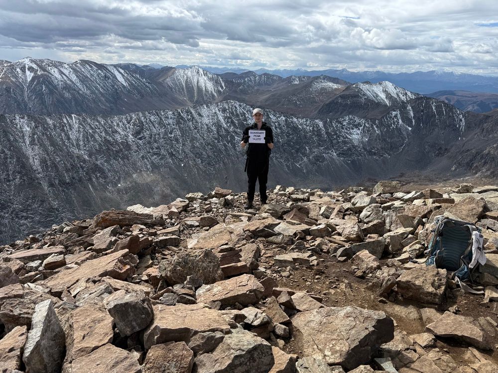 Neohew Duncan stands holding a sign on Quandary Peak, a 14er in Colorado. He is in a black top and bottom climbing gear, whir ball cap, standing on the top of a Rocky peak with two ranges of Rocky Mountains behind him. The peaks are snow topped but pine green covers most of the mountains. Duncan is the orignal Seasonal Nephew first posted 16 years ago in October as a Seasonal Nephew in Small Boy Massive Overgrown Pumpkin Season at the pumpkin patch visit. He is a good boy.  