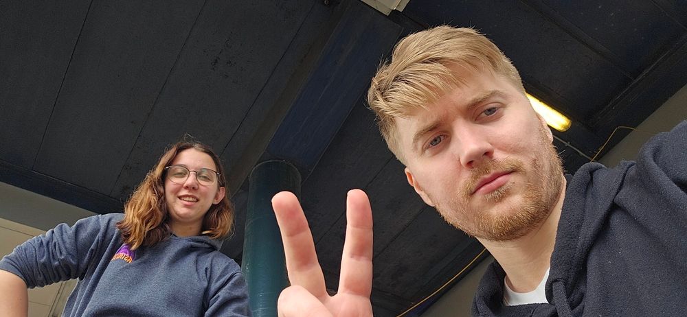 An image of two people in selfie style. On the left is one person looking confused at the photo being taken, the other person person on the right is looking dower and holding a peace sign towards the camera