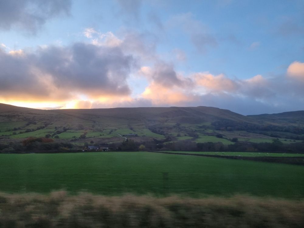 Sunrise over Edale, Peak District. Clouds tinged with gold in a pale blue sky float above a grassy ridge, with fields in foreground