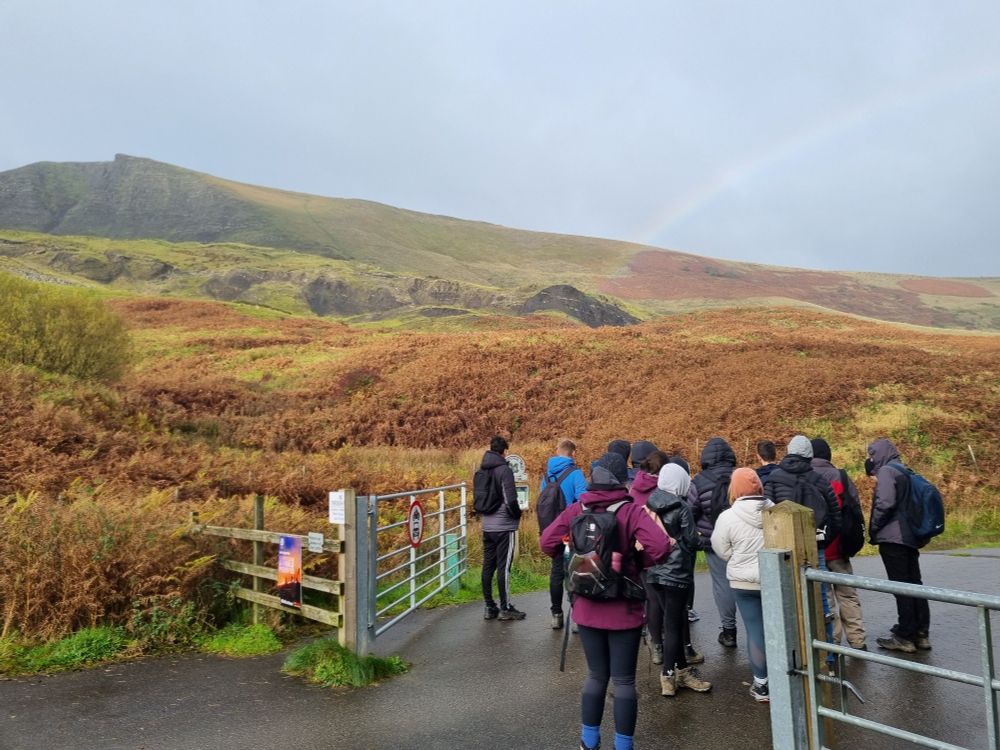 Students stand on tarmac track by gate, in front of bracken hummocky land of the Mam Tor landslide. A rainbow in the sky. Mam Tor escarpment in the background. Castleton, UK