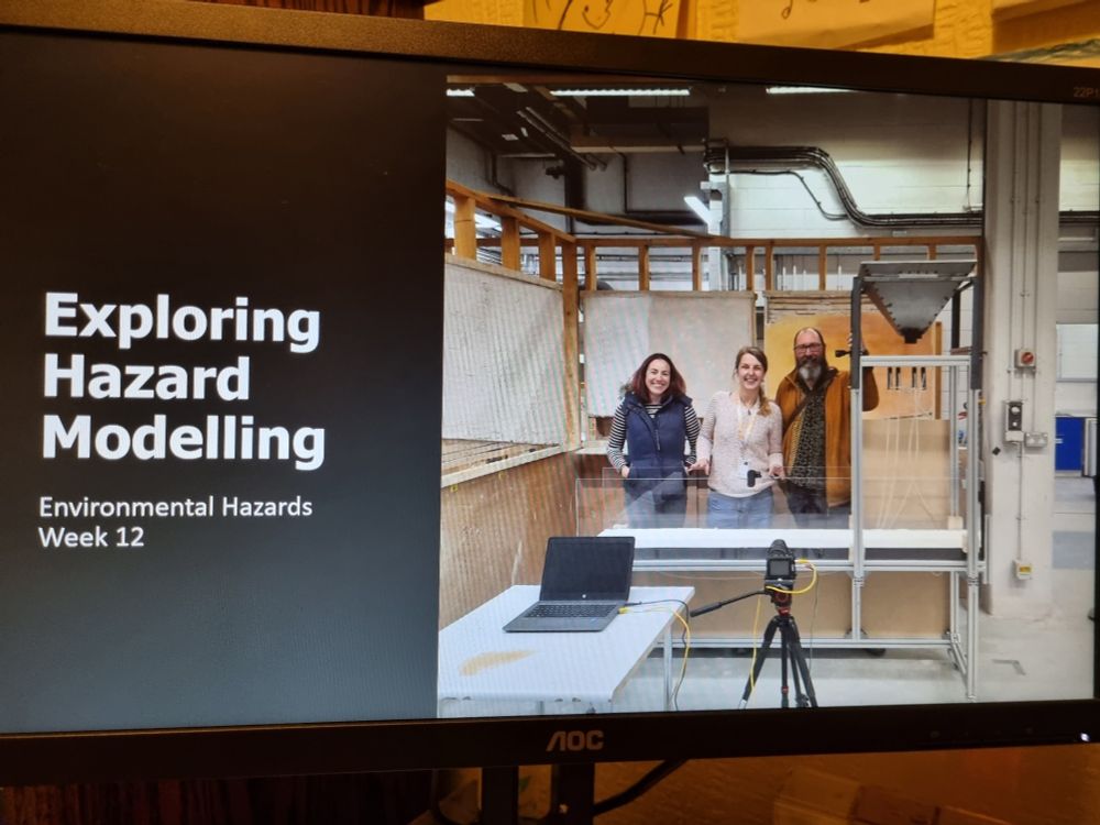A photo of a computer screen with the front page of a slide deck. It says "Exploring Hazard Modelling" next a photography of three scientists standing behind a experimental flume in a laboratory. The flume is made of a metal frame with a metal hopper and acrylic sides. A computer and camera are also in the image, in front of the flume.