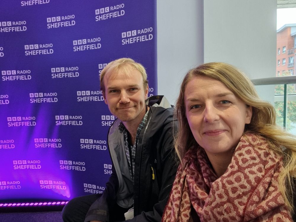 A woman with long hair and. Pink scarf sits in front of a man with short hair and a black coat. Behind them is a purple background with "BBC Radio Sheffield" written on it. The two lecturers are smiling nervously at the camera.
