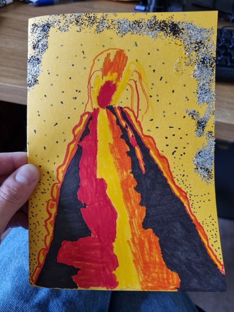 A yellow card with a drawing of a volcano. The volcano is black with red, yellow and orange lava shapes added, onckuding a red erupting fountain, and the sky above the volcano is filled with silver glitter, and black dots