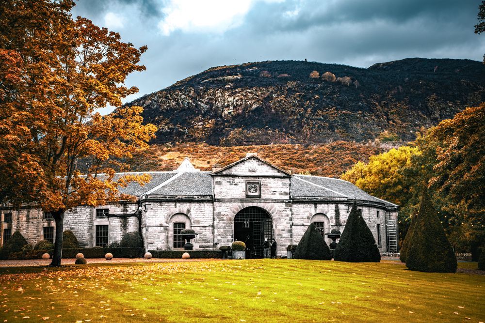 A striking, low-angle photograph of the historic Prestonfield House stables and courtyard, set against a dramatically contrasting backdrop. The single-story, stone building has a central arched entrance and a slate roof, and is flanked by clipped conical hedges and small topiaries. The foreground features a vibrant green lawn scattered with golden autumn leaves and a large tree with brilliant orange and yellow foliage on the left. Directly behind the building, the imposing, dark, and rugged slopes of Arthur's Seat dominate the upper half of the frame under a brooding, grey, and stormy sky. The surrounding trees on the hillside display the warm, rich colors of late autumn.