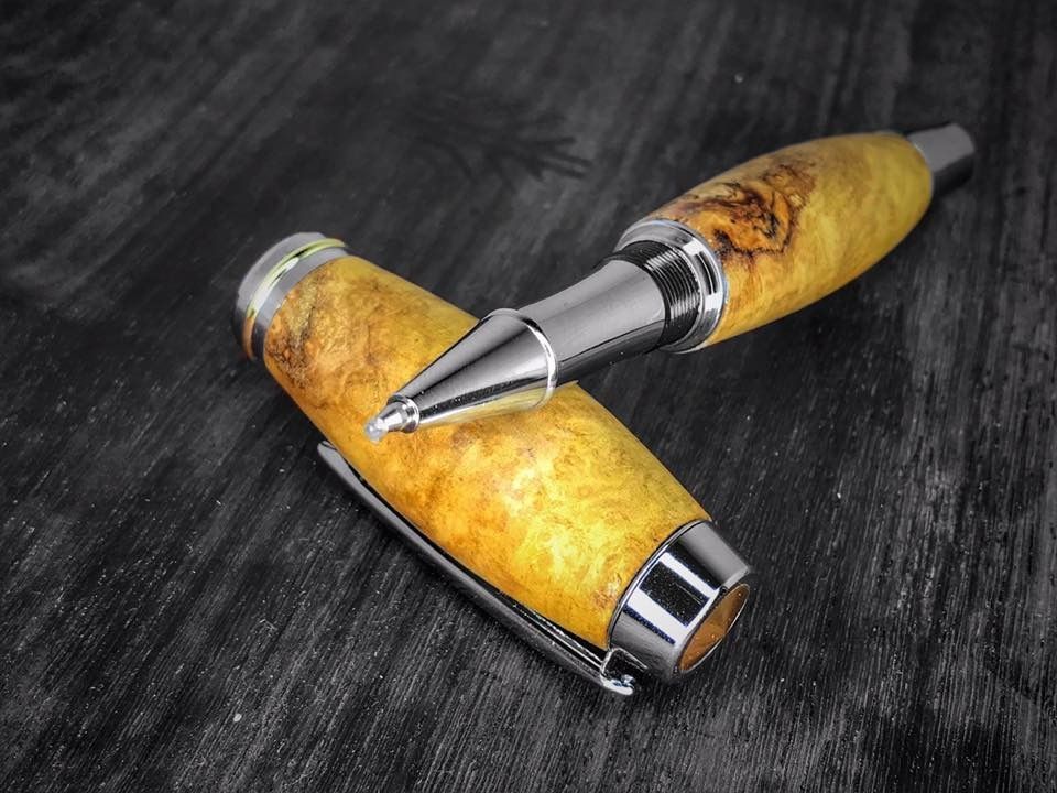 Close up shot of a handmade wood pen and its cap resting on a black wood surface. The pen itself is a silver color with a yellow wood grain. The cap is also yellow wood grain with a silver pen clip and silver rings around the cap.