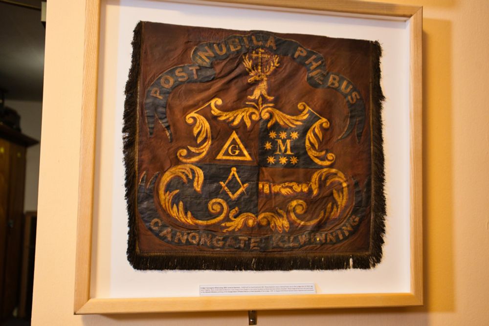 
The image shows a framed, old, textile banner or square hanging on a wall. The item is dark brown and appears to be made of velvet or a similar material. It is decorated with gold-colored embroidery.

The central design is a shield divided into four sections. The top left quarter has a "G" and a compass with a square, symbols often associated with Freemasonry. The top right quarter has what looks like a crown and three stars. The bottom two quarters are not as clear but appear to have swirling, ornate patterns.

Above the shield is a crown-like crest. Below the shield, a banner reads "CANONGATE KILWINNING".

Above the entire design, an arching banner reads "POST NUBILA PHOEBUS". The fringe of the textile is dark and appears to be worn. The item is protected behind glass in a light wooden frame.