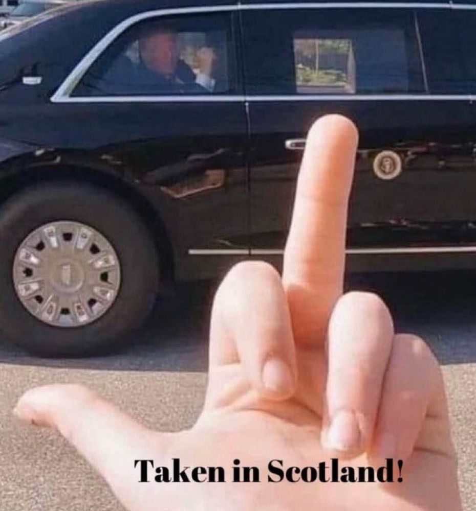 
A slightly blurry photo shows a person's hand in the foreground, giving a middle finger gesture. In the background, a large, black, armored car, likely a presidential limousine, is visible. A reflection in the car's window shows what appearsDonal Pedo Trump. Text at the bottom of the image reads "Taken in Scotland!".