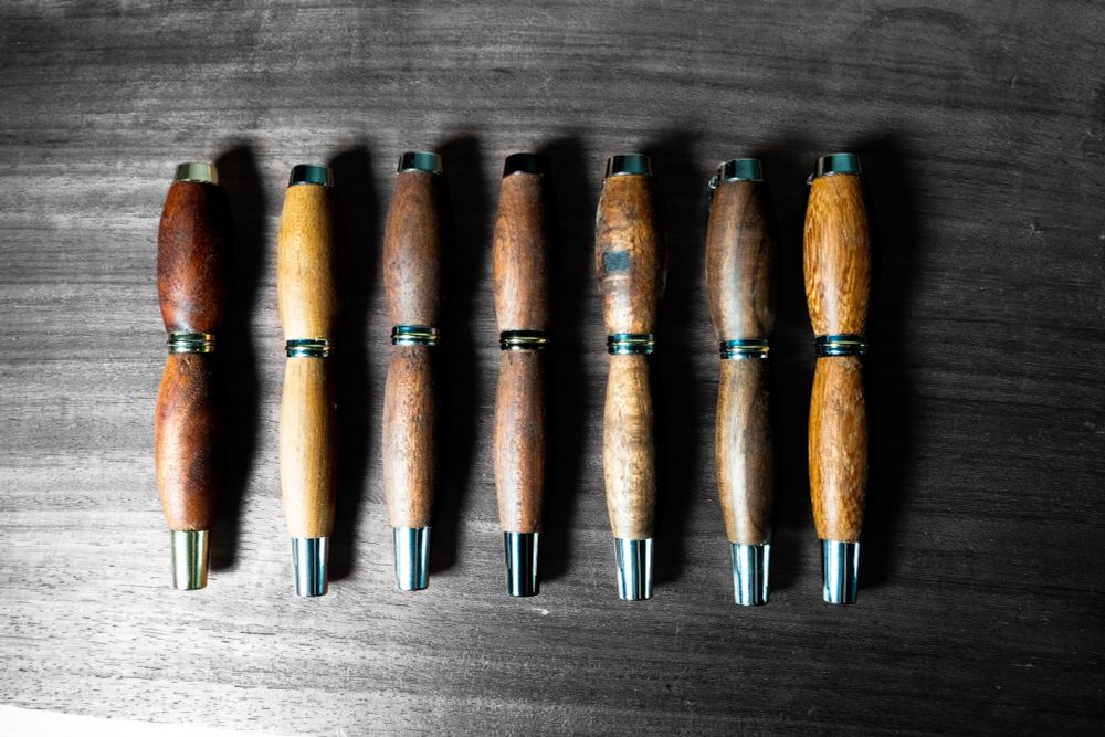A top-down shot shows eight wooden pens arranged horizontally on a dark, textured wooden surface. Each pen is handmade with a distinctive wood grain and a sleek, polished finish. They are evenly spaced in a single row across the center of the frame. The pens alternate in color, creating a pattern of light and dark wood. The first pen on the left is a rich, dark brown, followed by a lighter, golden-brown pen, and this alternating pattern continues across the row. Each pen features metallic accents at both ends and in the middle, likely gold or silver colored, which adds a touch of sophistication to their rustic design. The background surface is a deep, muted black with visible wood grain, providing a stark contrast that highlights the natural beauty and craftsmanship of the pens.