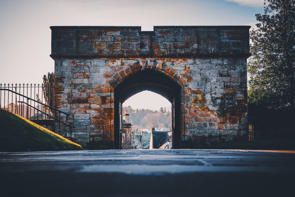 
A low-angle, wide shot of an old, weathered stone archway, likely a historic gatehouse, under a bright sky. The arch is built from large, rough-hewn blocks of light stone, heavily stained with patches of orange, brown, and black lichen/weathering. The archway frames a view of a grassy hill sloping downwards on the other side, leading to rooftops and trees in the distance under a slightly hazy sun. The foreground is dominated by a dark, blurred pavement or road surface. To the left, a grassy slope meets a black metal railing and a short flight of steps leading up. The overall mood is historic and atmospheric, with warm light highlighting the stonework's texture.