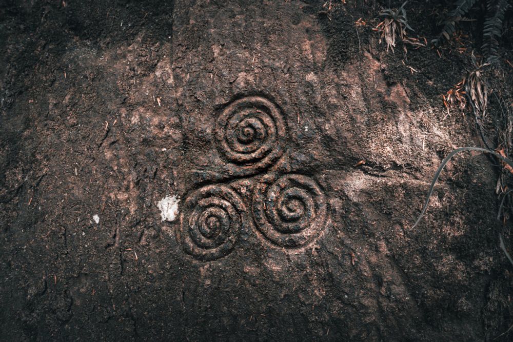 A close-up shot of a dark and textured stone surface featuring a carved triskelion, which is a motif consisting of three interlocked spirals, likely found in Roslin Glen Country Park near Wallace cave. The stone's surface is rough and weathered, with small bits of debris scattered around the carving. The triskelion itself is deeply etched into the stone, with the spirals evenly spaced and connected at the center. To the right of the stone, some ferns and other foliage add a touch of nature to the scene. The overall tone of the image is earthy, highlighting the natural and historical aspects of the location.