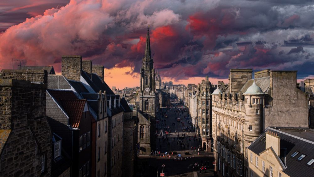 A dramatic view of Edinburgh’s Royal Mile with a moody sky filled with deep red and dark grey clouds. The historic street stretches toward the horizon, lined with old stone buildings, leading to St. Giles’ Cathedral and Edinburgh Castle in the distance. The sunset casts a warm glow, contrasting with the stormy atmosphere.