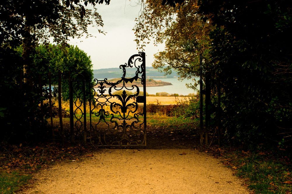 A striking, centered photograph of a partially open, highly ornate black wrought-iron gate framing a scenic view, set within a shadowed border of dense autumn foliage. The path leading up to the gate is a wide, light-brown gravel track. Beyond the gate, the bright landscape opens up to a sunlit, golden-hued field that slopes down to a wide, calm estuary or bay of water. A distant, wooded shoreline forms the far bank under a muted, overcast sky. The image has a dramatic contrast, emphasizing the dark, intricate metalwork of the gate against the bright vista. The location is an estuary view near Lauriston Castle in Edinburgh. The scene captures a feeling of stepping from shadow into light and a hidden, majestic view.
