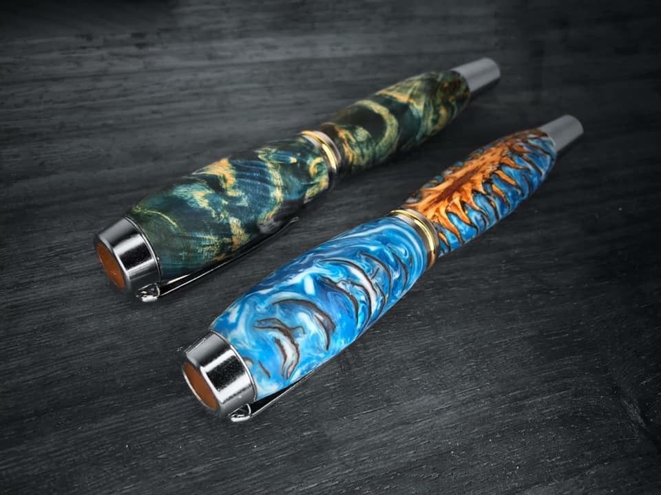 Two handcrafted pens rest diagonally on a dark gray wooden surface. The pen closer to the top of the image has a swirling pattern of blue, green, and gold colors, resembling a stormy sea. The second pen, closer to the bottom, features a striking combination of light blue and white swirling patterns, complemented by a section of raised, spiky textures in a natural wood color that resembles a pinecone. Both pens have a sleek, silver-colored clip and band, adding a touch of elegance to their unique designs.