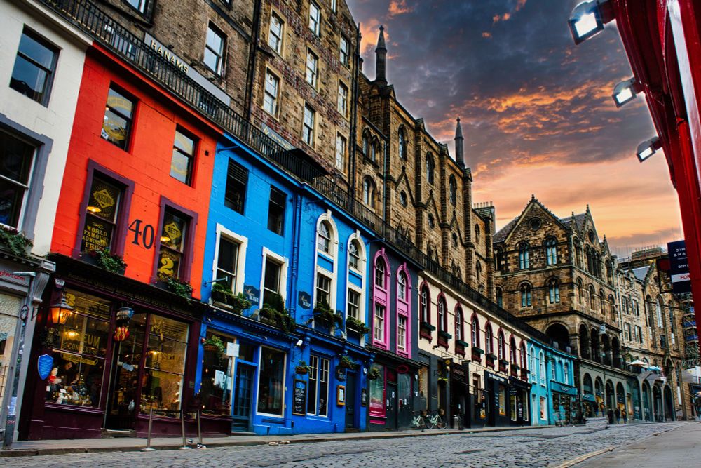 A vibrant photo of Victoria Street in Edinburgh, lined with colorful shopfronts and historic stone buildings under a dramatic sunset sky.
