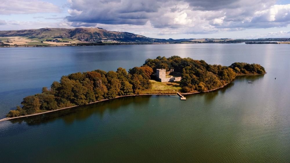 Lochleven Castle A high-angle, long shot shows a ruined castle on a heavily-wooded island in the middle of a lake. The castle, situated in the middle of the island, consists of a square tower and a ruined wall. It is surrounded by a grassy field, and a small wooden dock leads out into the water from the shore. The island itself is covered in trees that are turning yellow and brown for autumn. A small strip of land extends out from the island into the lake at the left edge of the shot. The lake has some ripples, and the shoreline of the opposite shore is visible. Behind the shore are brown and green hills and some scattered houses and buildings. The sky is overcast with some white clouds and some dark clouds.