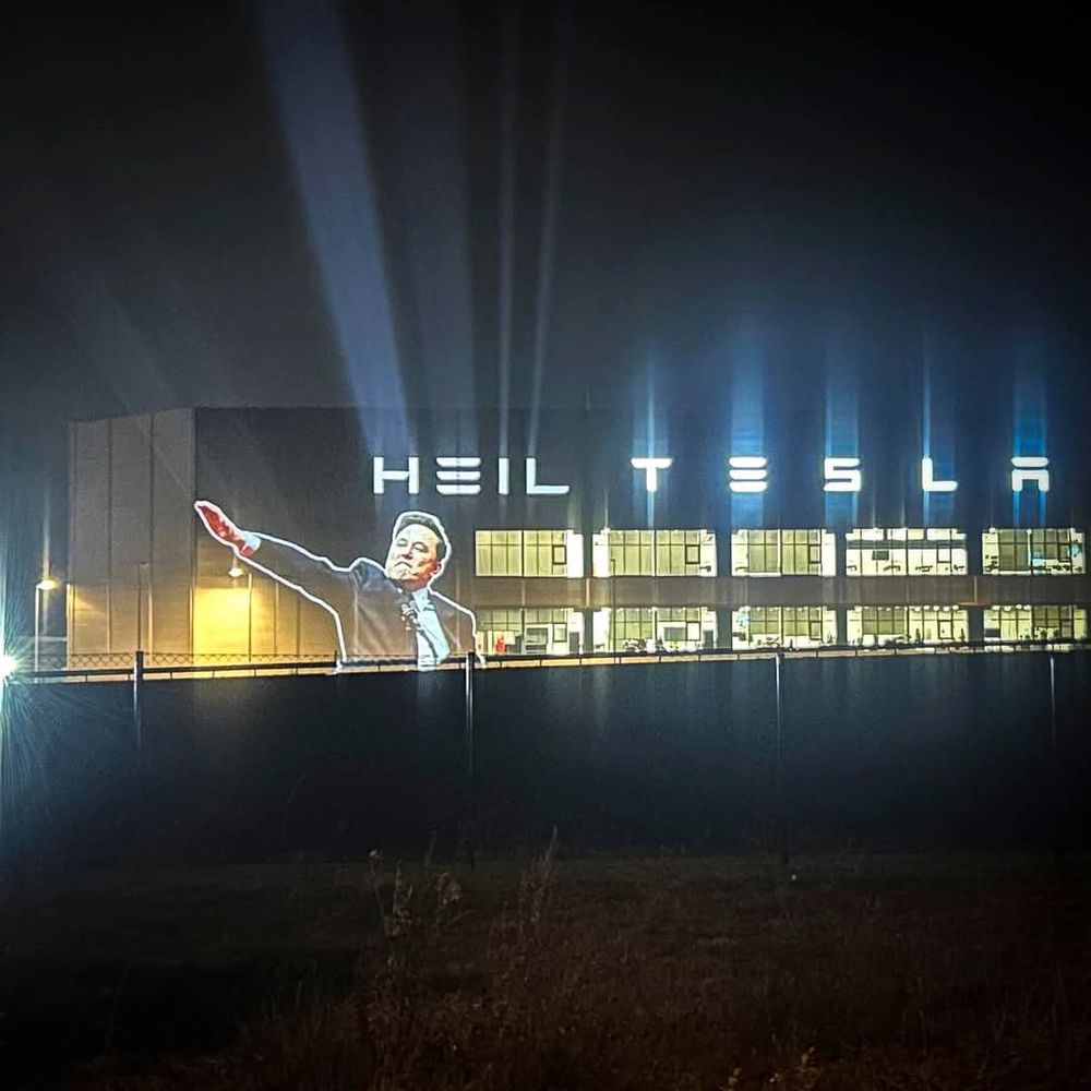 A Tesla factory at night with a projection of Elon Musk giving a Nazi salute and the words “Heil Tesla” lit up behind him on the building.