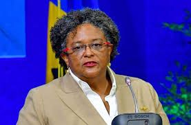 A woman with short dark grey hair and a serious expression speaking at a microphone. She is wearing a beige jacket, white shirt, and glasses with dark pink frames; against a bright blue background.