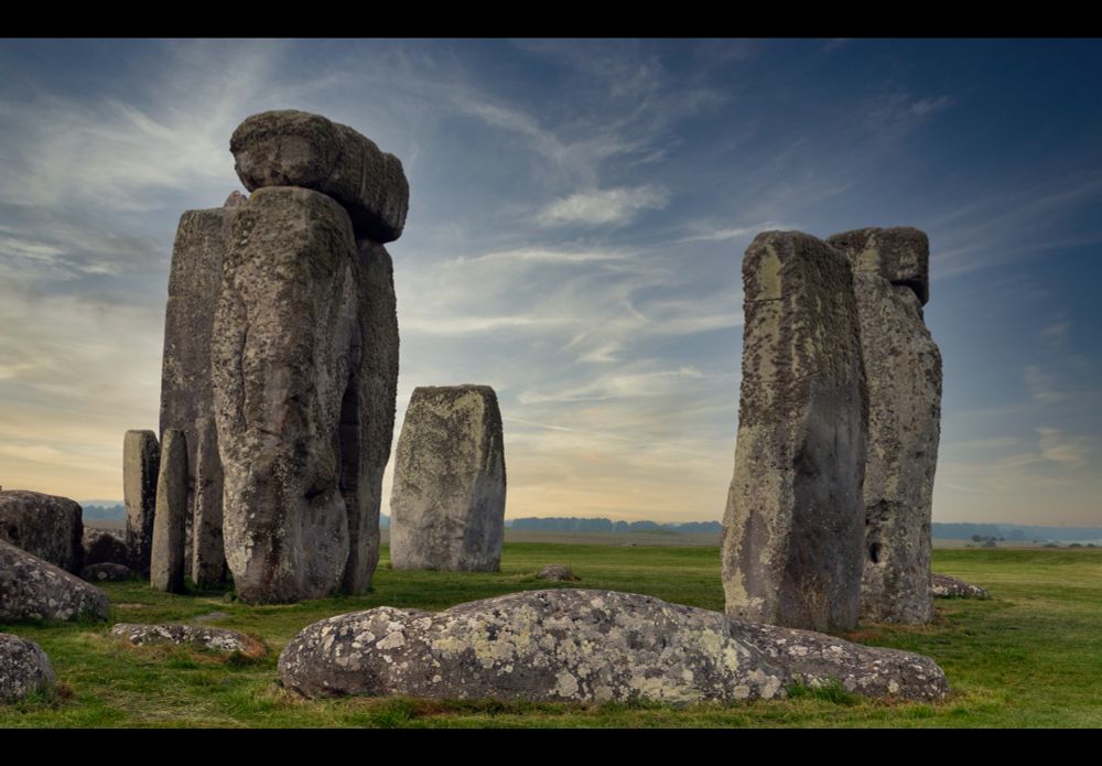 this is an image of a part of Stonehenge. It is not the entire site. Just a small section of it. The sky is blue with thin wispy clouds. The sun is setting.