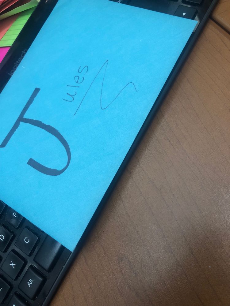 A blue envelope with “Jules” written on it is sitting on a black keyboard, on a brown faux-wood desk.