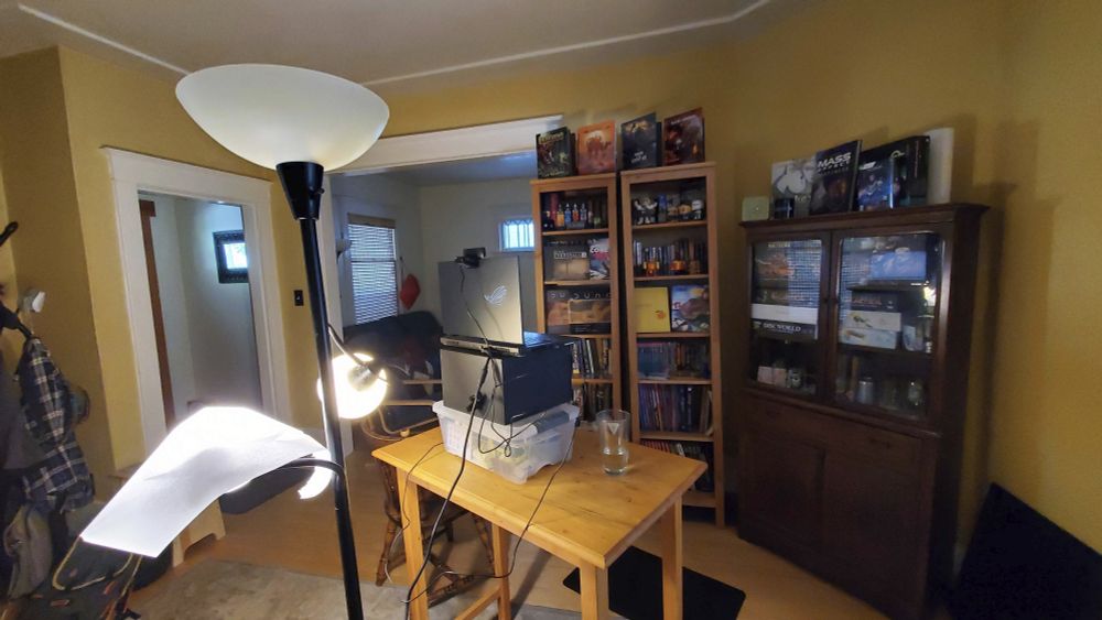 A cluttered living room with diffused lighting. a laptop area points at bookshelves filled with books, board games, collectibles, and other geeky stuff. 