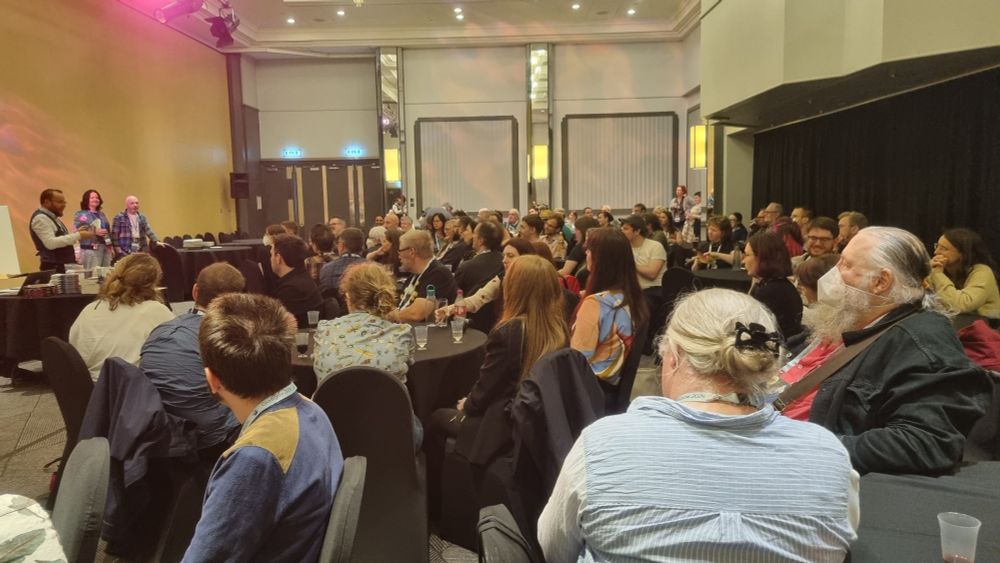 At the Gallus launch on 10th August at Glasgow Worldcon. In the foreground, a full room of people sat at round tables with black tablecloths. At the side of the room, editors Brian M. Milton, E.M. Faulds and Neil Williamson introduce the book in front of a table of copies." title="At the Gallus launch on 10th August at Glasgow Worldcon. In the foreground, a full room of people sat at round tables with black tablecloths. At the side of the room, editors Brian M. Milton, E.M. Faulds and Neil Williamson introduce the book in front of a table of copies.