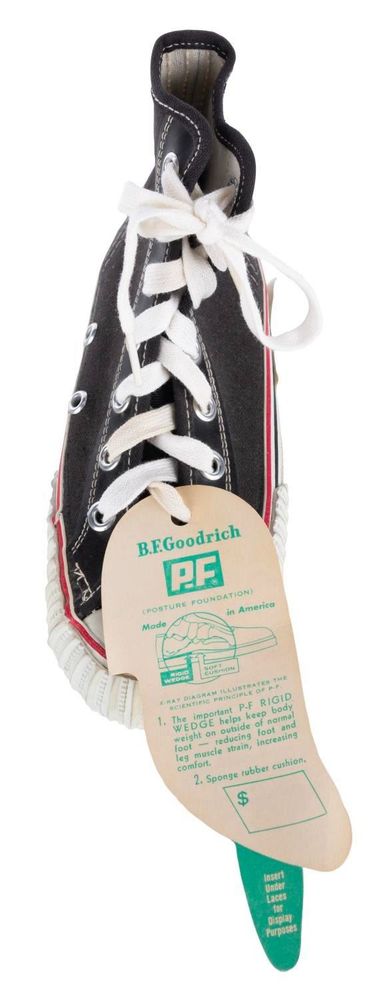 Close up of the actual sneaker that rests on the base of the display. It is black with white shoelaces and rubber soles, with red striping around the edges. A large paper tag hangs from the laces near the toe. It has a diagram of the shoe and describes how it is constructed, with a large empty box at the bottom to fill in the price. It states that the shoes are made in America, and reads:
“1. The important P.F RIGID WEDGE helps keep body weight on outside of normal foot
- reducing foot and leg muscle strain, increasing comfort.
2. Sponge rubber cushion.”
They are made by B.F. Goodrich, and the PF stands for Posture Foundation.