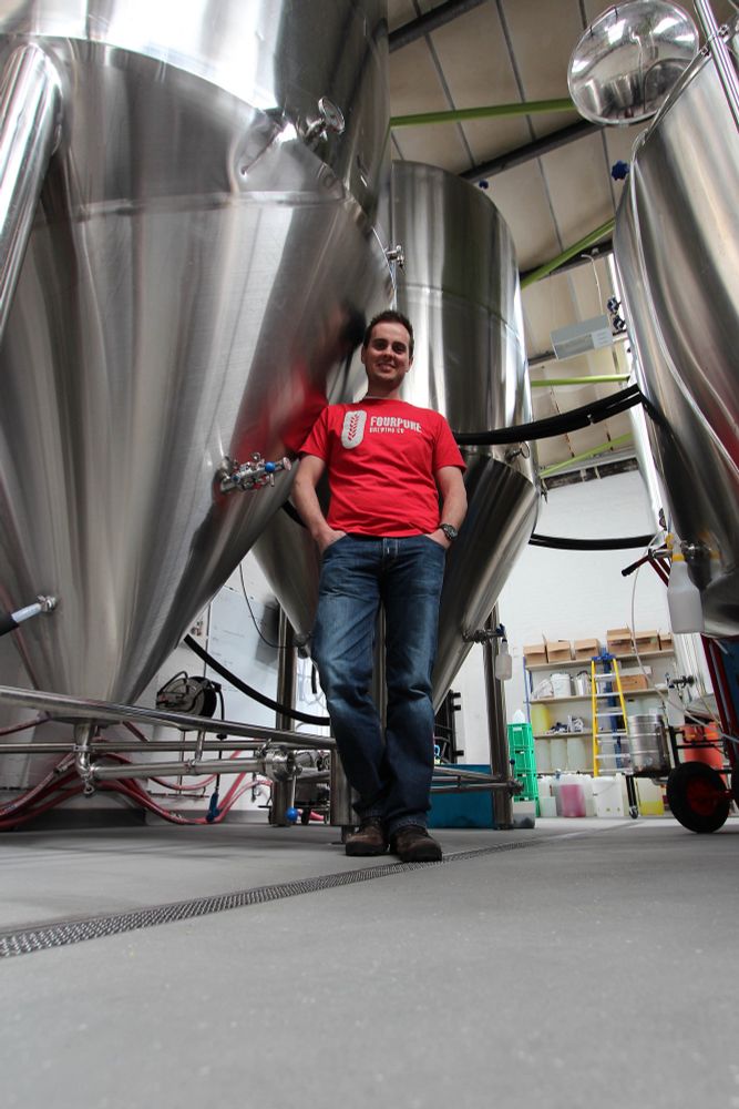 A man in a red tee shirt and jeans stands in front of a stainless steel conical fermentation tank in a micro brewery.