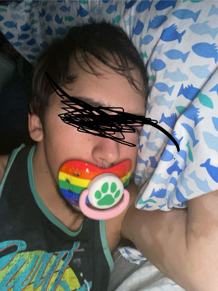 My head laying on a pillow with a big pacifier with a rainbow shield, pink ring, and a white button adorning a green puppy paw in my mouth. 