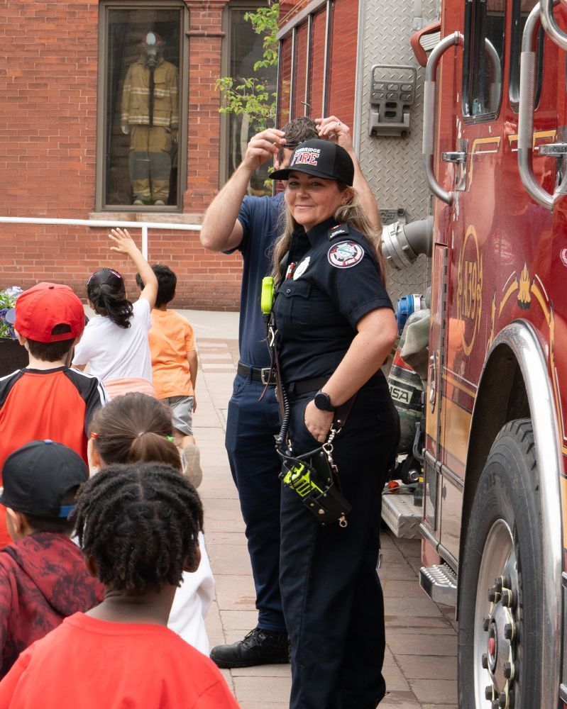 Two firefighters from the City of Cambridge interacting with a group of children near a fire truck.
