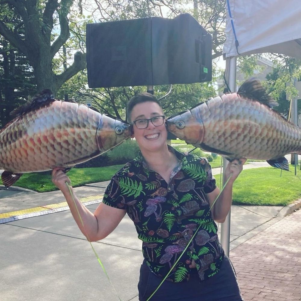 El, wearing a delighted expression and a shirt printed with mushrooms and leaves, poses with two carp-shaped balloons at the Pleasant Peninsula art and activism festival in Grand Rapids MI.