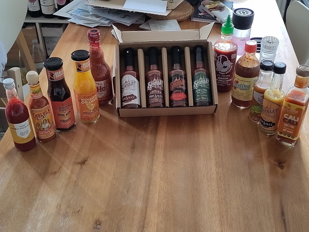 On a table are 14 different hot sauces and bbq glazes. These range from a custom one on the far left through staples like Frank's Hot sauce, and includes a number of small batch hot sauces from the Dublin hot sauce company Scarlet For Yer Ma.