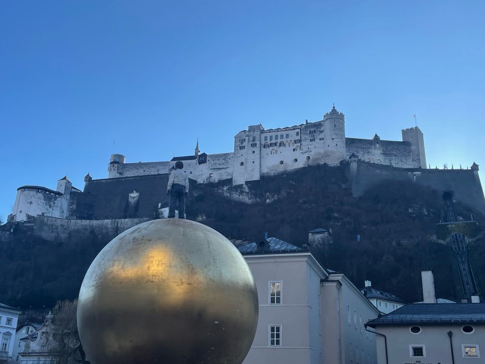 Fortress high on a hill with a golden ball sculpture in the foreground 